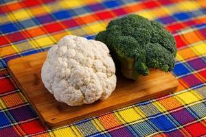 a head of broccoli and a head of cauliflower lie on a cutting board on the table with colorful towel underneath photo