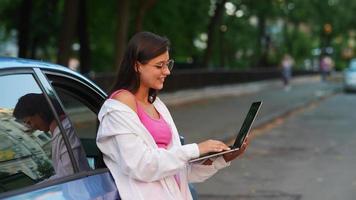 la mujer se apoya en el auto mientras sostiene la computadora portátil y escribe