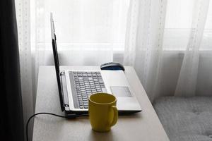 Home office concept. Work from home in living room. Laptop and cup of hot beverage on wooden table near sofa and window. photo