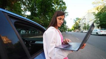 Woman leans on car while holding laptop and typing video