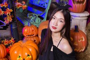 Asian woman wears ghost black coat and makes up as witch ghost while looks at the camera and there is a pumpkin head jack lantern placed on the side at night in outdoor setting against background.