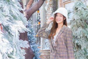 hermosa mujer asiática usa un sombrero blanco y un abrigo a cuadros mientras mira la cámara y se para al aire libre cerca de la nieve cubierta de árboles de navidad en el tema de año nuevo e invierno. foto