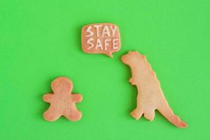 Homemade cookies in shapes of dinosaur and man with inscription - Stay safe - on green background, top view. Sweet shortbread with white glaze. Social distancing concept. photo
