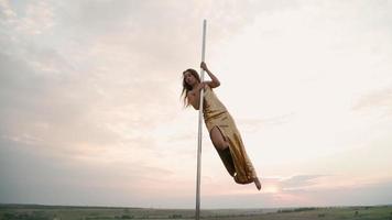 Beautiful athletic woman in a gold dress performs tricks on the pole against the background of sunset in the field. Beauty and body care. Female sports and fitness. Slow motion video