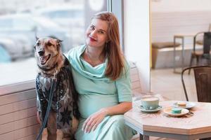 Beautiful young pregnant women is hugging her dog in cafe with cup of coffee and macaroons on a table. photo