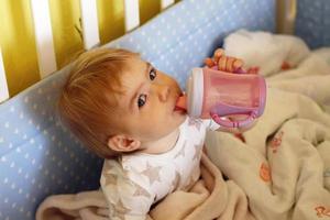 linda niña de un año con ojos azules y rubia está bebiendo agua de una botella después de despertarse por la mañana en su cama. foto