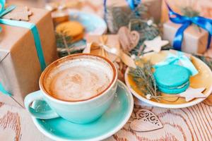 Blue cup with cappuccino, sweet macarons on blue and yellow saucers, with Christmas gifts, Christmas decorations and coniferous branches on wooden background, top view. photo