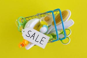 Flat lay on shopping basket with baby care items - scissors, hairbrushes, pacifiers, thermometer, cotton pads, pacifier holders and nasal aspirator - on yellow background. photo