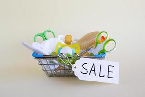 Shopping basket with baby care items - scissors, hairbrushes, pacifiers, thermometer, cotton pads, pacifier holders and nasal aspirator - on a beige background with paper price tag with text 'Sale'. photo