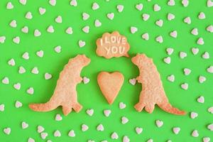 Homemade shortbread cookies decorated white glaze. Two dinosaurs with callout cloud with text - I love you - on green background with heart sprinkles, top view photo