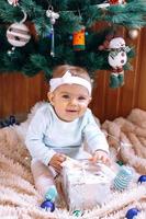 Smiling little girl is sitting near Christmas tree on a beige plush plaid with Christmas decorations and lights and try to open Christmas gift. photo