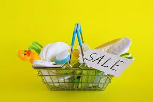 Shopping basket with baby care items - scissors, hairbrushes, pacifiers, thermometer, cotton pads, pacifier holders and nasal aspirator - on yellow background with paper price tag with text 'Sale'. photo