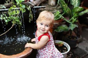 niña sonriente con cabello rubio y ojos azules en sundress está de pie en el jardín con macetas y cascada. foto