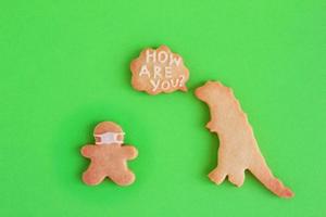 Homemade shortbread cookies in shapes of dinosaur and man in face mask and with callout cloud with text - How are you - on green background. Social distancing concept. photo