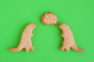 Two homemade cookies in shapes of dinosaurs with inscription - Stay home - on green background, top view. Sweet shortbread with white glaze. Social distancing concept. photo