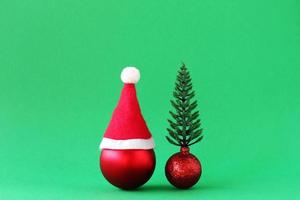 Red Christmas ball with Santa Claus red hat and shiny red ball with artificial Christmas tree on green background with copy space. Minimal Christmas and New Year concept. photo