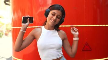 une jeune femme chante et danse devant un mur rouge tout en portant des écouteurs et en chantant dans un smartphone video