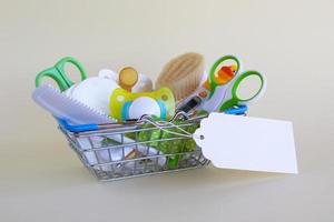 Shopping basket with baby care items - scissors, hairbrushes, pacifiers, thermometer, cotton pads, pacifier holders and nasal aspirator - on a beige background with blank paper price tag. photo