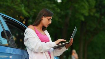 Woman leans on car while holding laptop and typing video