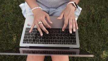 vista de arriba hacia abajo de una mujer escribiendo en un teclado portátil mientras está sentada afuera en el césped