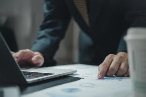 Businessman hand pointing paperwork chart report analysis statistic and document financial graph with computer laptop at office.Business investment marketing on desk in company. photo