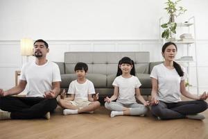 feliz familia tailandesa asiática, padres e hijos se sientan en el piso de la sala de estar, cierran los ojos juntos, meditan tranquilamente en posición de loto y practican yoga saludable, fin de semana de estilo de vida doméstico. foto