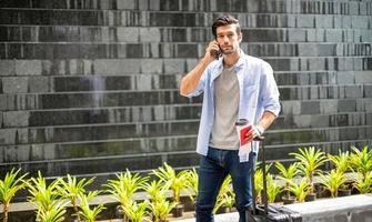 Young caucasian man using smartphone talking and pulling the suitcase waiting his friend for travel together. photo