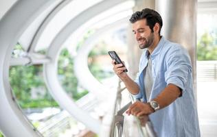 Young caucasian man using smart phone and smiling while waiting his friend at the outside. The man holding and using cellphone for searching data and social media on internet. photo
