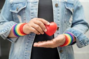 Asian lady wearing rainbow flag wristbands and hold red heart, symbol of LGBT pride month celebrate annual in June social of gay, lesbian, bisexual, transgender, human rights. photo
