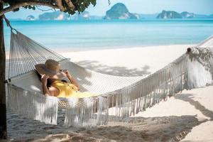 feliz mujer viajera se relaja en una hamaca en la playa del paraíso. mujer turista vestida de amarillo descansa cerca del mar tropical. concepto de vacaciones, viajes, verano, pasión por los viajes y vacaciones foto