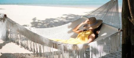 feliz mujer viajera se relaja en una hamaca en la playa del paraíso. mujer turista vestida de amarillo descansa cerca del mar tropical. concepto de vacaciones, viajes, verano, pasión por los viajes y vacaciones foto