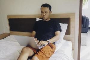 Happy young man working from the bed. Put the laptop on the bed and type on the keyboard. photo