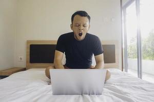 A sleepy young man yawning while checking laptop on the bed. photo