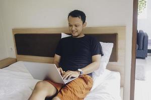 Smiling young man working from the bed. Hold the laptop on the knees and type on the keyboard. photo