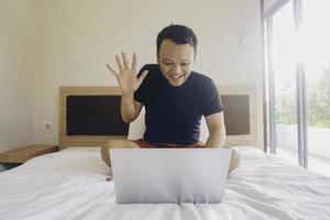 Smiling young Asian man works remotely behind a laptop at home, sitting in bed photo