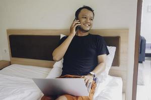 Happy young man working from the bed. Hold the laptop on the knees and talking on the phone. photo