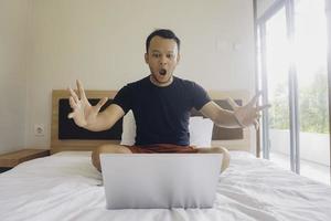 Shocked young man checking on his laptop while sitting in bed photo