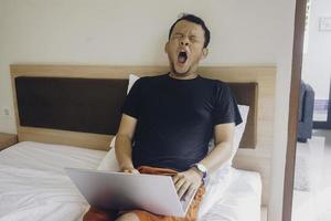 A sleepy young man yawning while checking laptop on the bed. photo