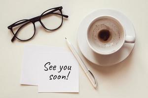 See you soon note, empty cup with coffee leftovers, eyeglasses and pen on white desk photo