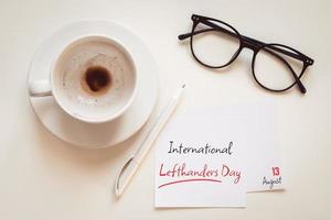 International lefthanders day greeting on August 13, desktop flat lay with coffee cup, pen, eyeglasses and paper note photo