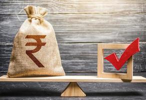 Red vote tick and a indian rupee money bag on scales. Estimating cost of making a decision and consequences in the future. Corruption risks. Concept of lobbying for decisions and laws. photo