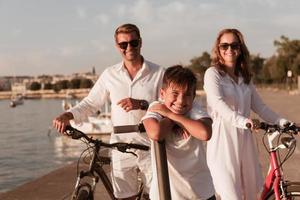 familia feliz disfrutando juntos de una hermosa mañana junto al mar, padres montando en bicicleta y su hijo montando una scooter eléctrica. enfoque selectivo foto