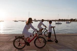 familia feliz disfrutando juntos de una hermosa mañana junto al mar, padres montando en bicicleta y su hijo montando una scooter eléctrica. enfoque selectivo foto