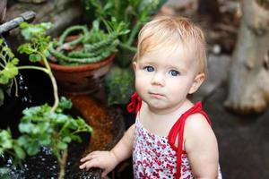 Portrait of little girl with blond hair and blue eyes in sundress in garden with pot plants and waterfall. photo