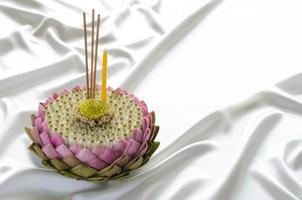 krathong de pétalos de loto rosa para tailandia el festival loy krathong decora con su polen, flor de corona, palo de incienso y vela sobre fondo de tela blanca suave y ondulada. foto