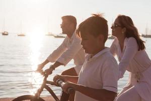 Happy family enjoying a beautiful morning by the sea together, parents riding a bike and their son riding an electric scooter. Selective focus photo