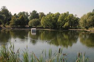 White wooden floating duck house in the centre of the pond in public park. Bird sanctuary in artificial lake. Spring nest. Summer green view. Wildlife nature reserve in river water. Waterfowl shelter photo