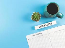 plano de libro de seguimiento de hábitos con calendario de madera de diciembre, taza azul de café negro y maceta suculenta sobre fondo azul con espacio para copiar. foto