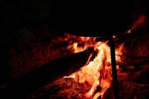 Food cooking on a bonfire at night. Firewood solid fuel for cook at campfire. Fire with orange flame on dark background. Burning wood for heating energy. Dinner at camping. Fire to keep warm at camp. photo