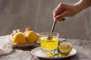 Ginger tea with lemon, ginger root and rosemarry on wooden background. Small glass transparent pitcher with hot drink. Seasonal beverages. Shallow DOF, selective focus, focus on top of pitcher. photo